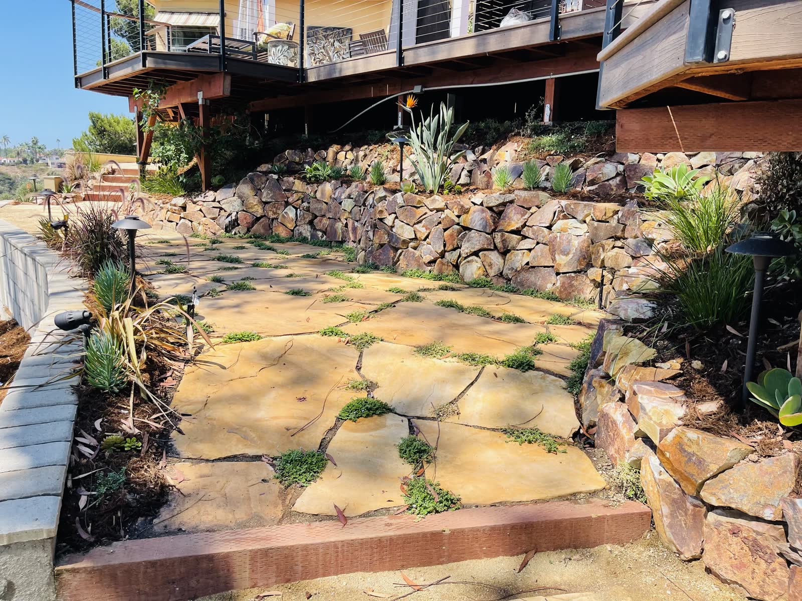 Flagstone patio with planted joints below a natural boulder retaining wall and an elevated deck
