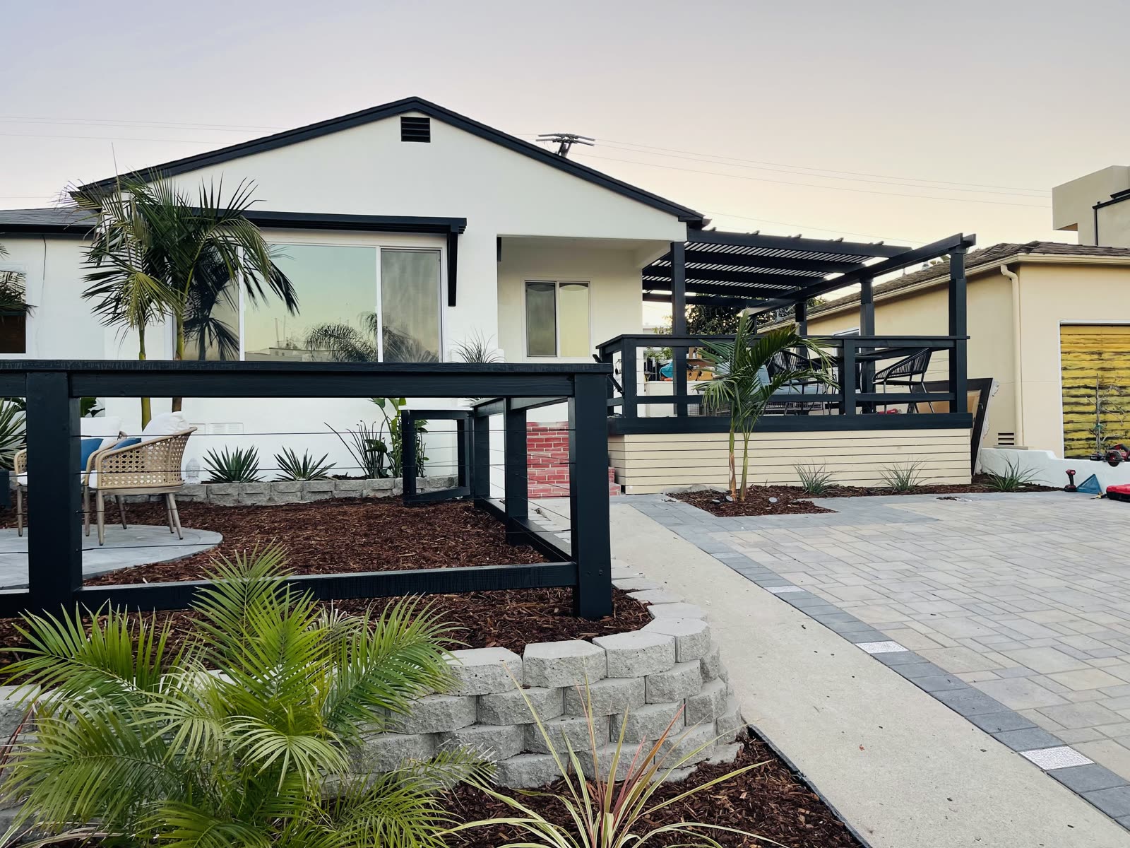 Modern front yard with a black pergola, cable railing, paver driveway, and fresh planting beds